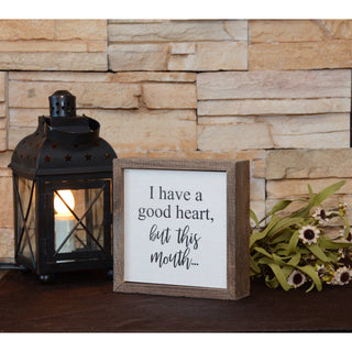 Decorative sign with text next to a lantern and flowers against a stone wall.